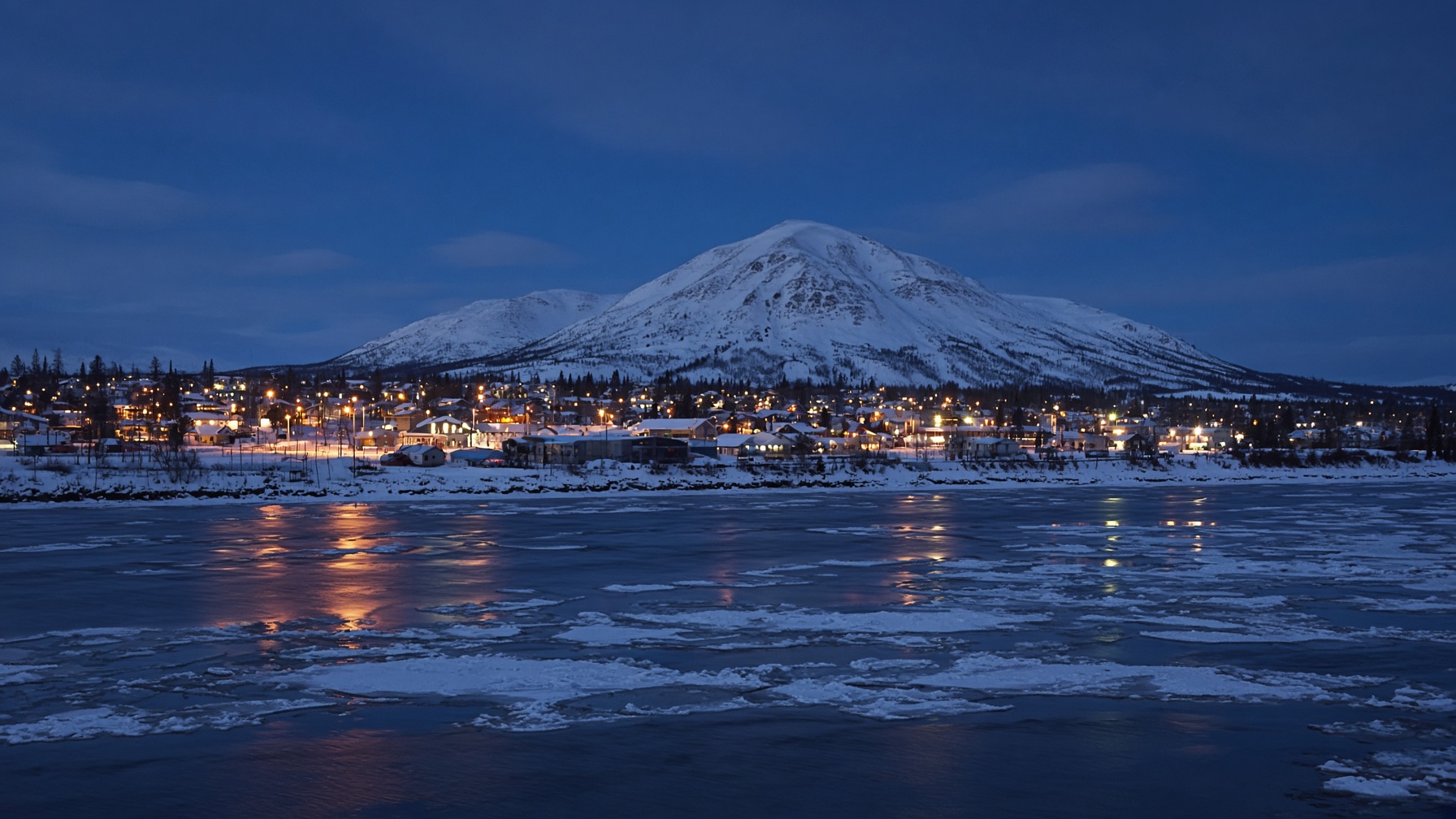 Winter dusk over a northern river town with lights reflecting on ice and a mountain beyond.